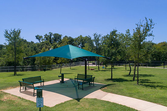 Blue pyramid shade installation at a community park, the shaded area has seating, and the green fabric is creating blue shade on the ground.